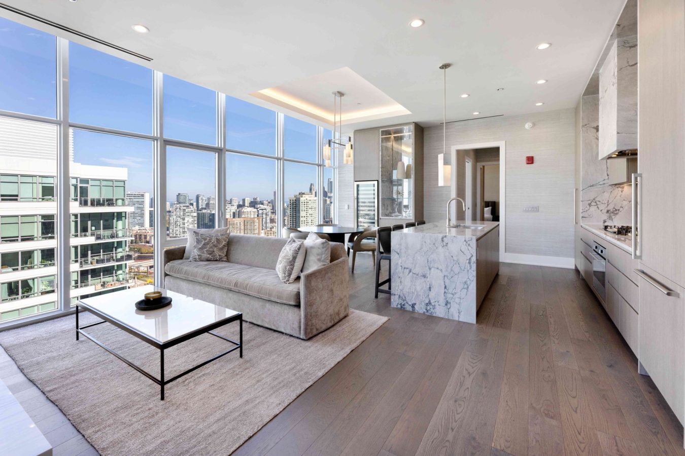 Modern apartment interior with floor-to-ceiling windows, city skyline view, a gray sofa, white marble kitchen island, sleek cabinetry, built-in appliances, and a light wood floor.