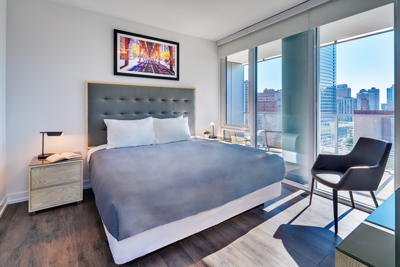 Modern bedroom with large windows, city view, a gray upholstered bed with white pillows, two bedside tables with lamps, a black chair, and a framed cityscape artwork above the bed. Sunlight brightens the room.