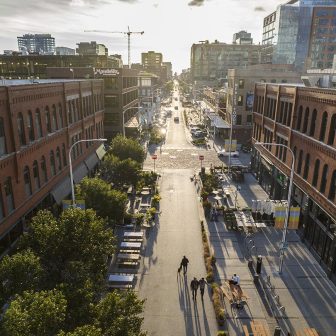 Aerial view of a city street lined with red brick buildings, outdoor seating areas, and trees; a few people walk along the street as sunlight casts long shadows. Modern buildings and a crane are visible in the background.