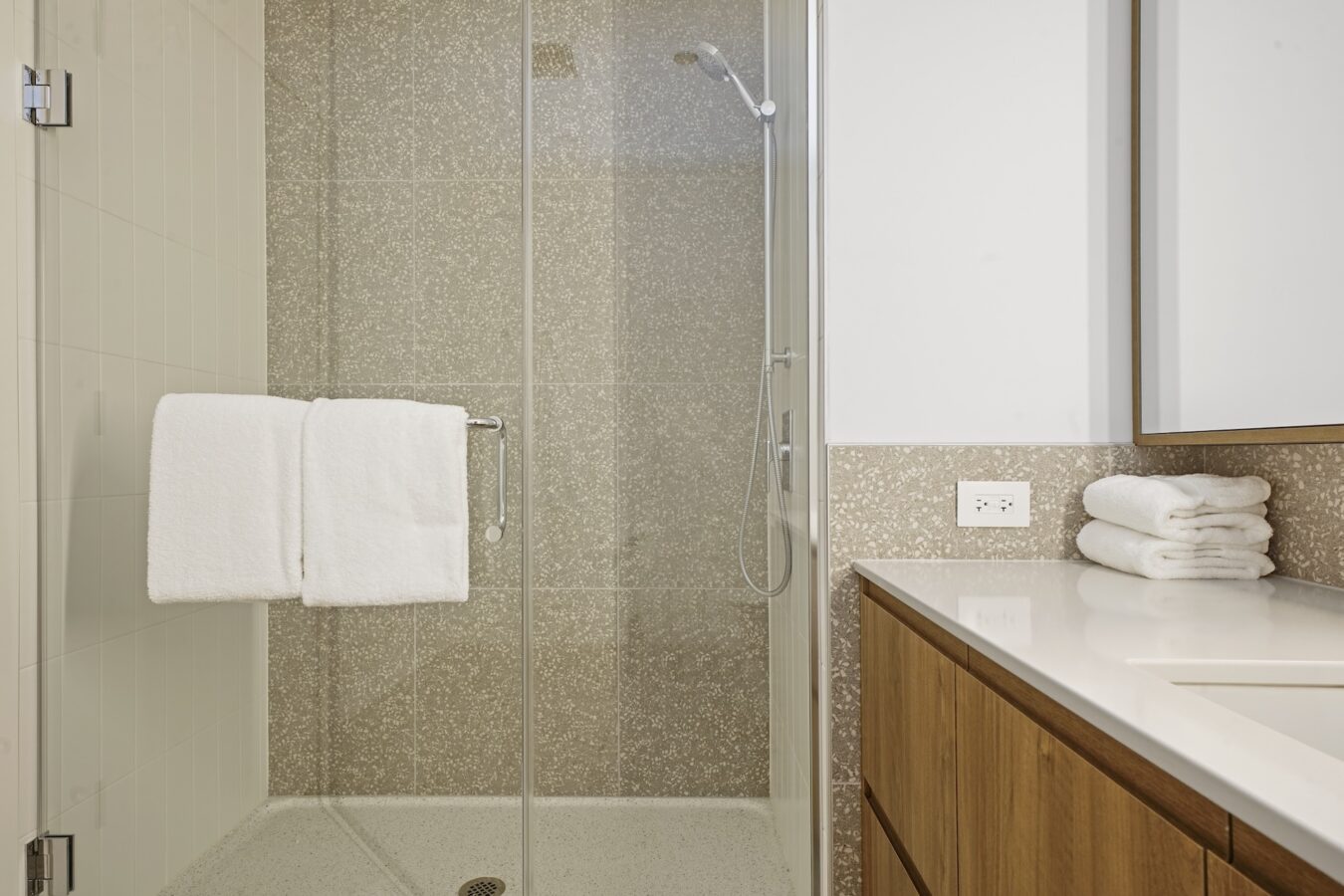Modern bathroom with a glass shower enclosure, beige tiled walls, a showerhead, a towel hanging on the shower door, wooden vanity with a white countertop, and neatly folded towels beside the sink.