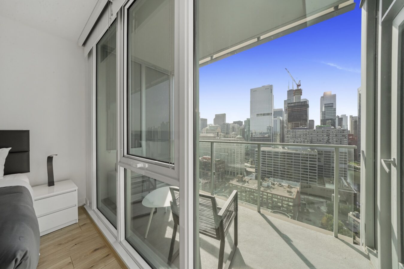 A modern bedroom with floor-to-ceiling glass doors opens to a balcony overlooking a city skyline with tall buildings, blue sky, and a construction crane in the distance.