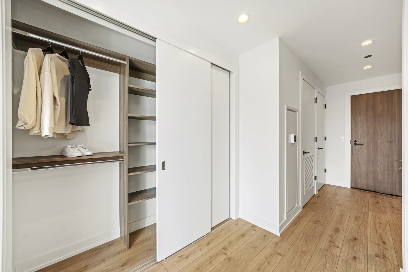 Minimalist hallway with a wooden floor, an open closet featuring hanging clothes and shelves, and a closed wooden entrance door. The closet has sliding white doors and built-in shelving.