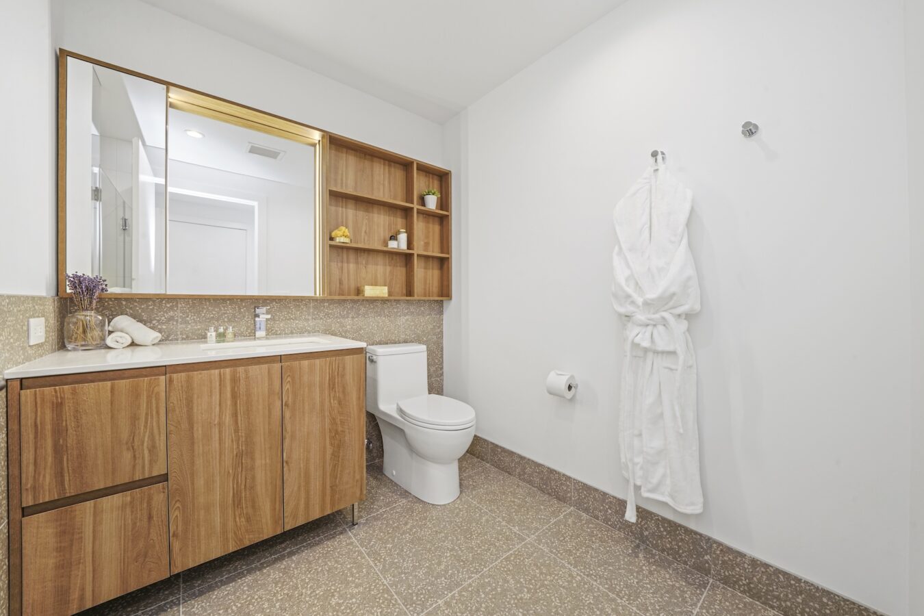 Modern bathroom with a wooden vanity, large mirror, white toilet, towel rack with a hanging white robe, open shelves with decor, and neutral tile flooring and walls.