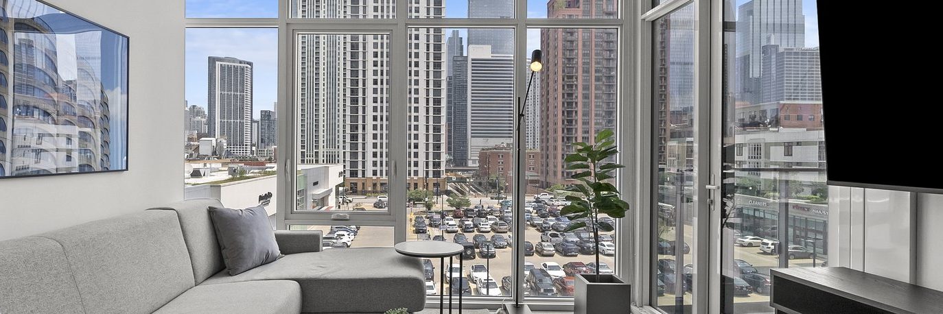 Modern living room with a gray sectional sofa and large floor-to-ceiling windows overlooking a cityscape with tall buildings and a parking lot below. A small table and a potted plant are near the window.