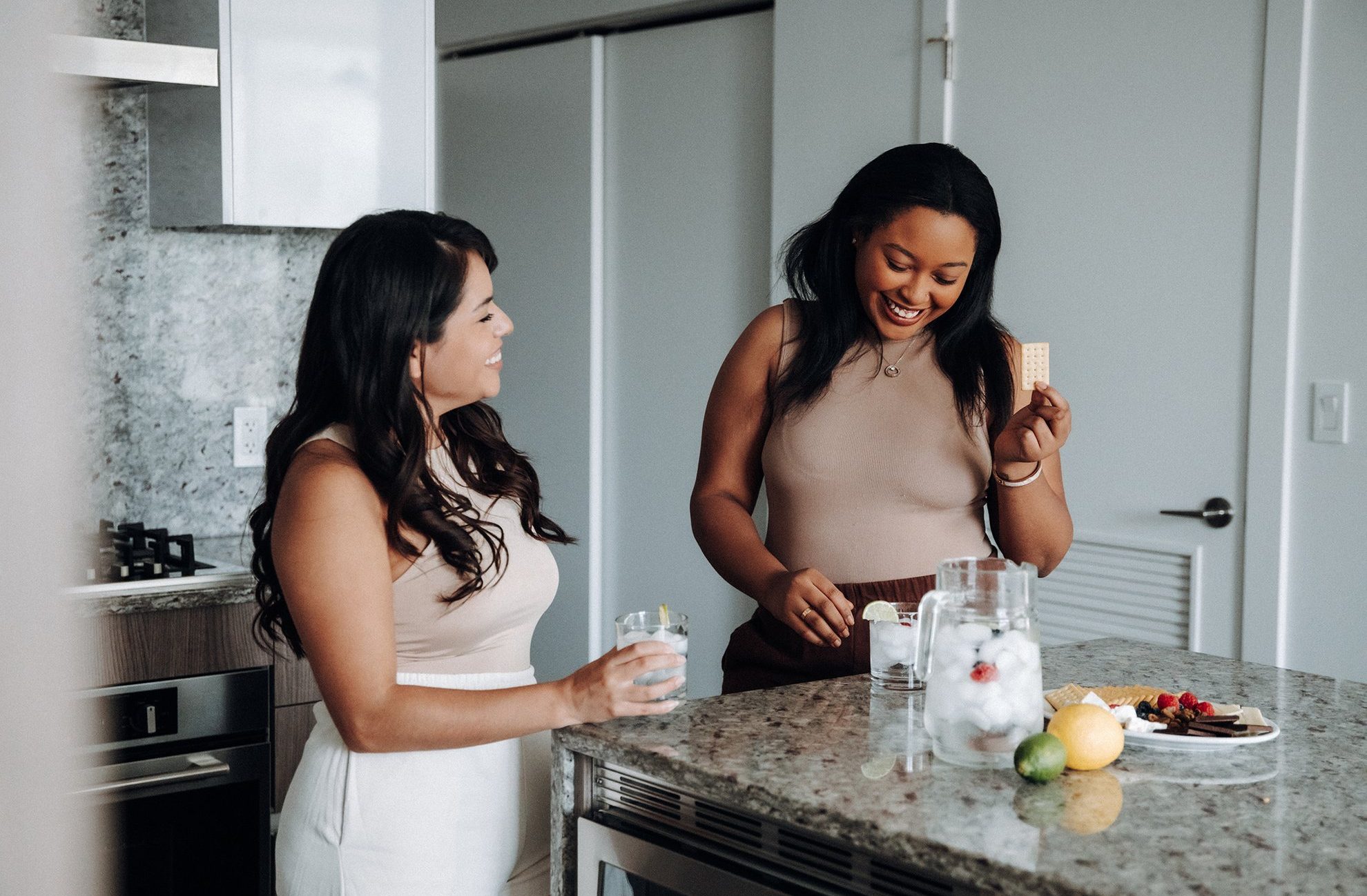 Two women stand in a modern kitchen, smiling and talking. One holds a glass of water with lemon, while the other holds a cracker by a bowl of fruit and ice water on the countertop.