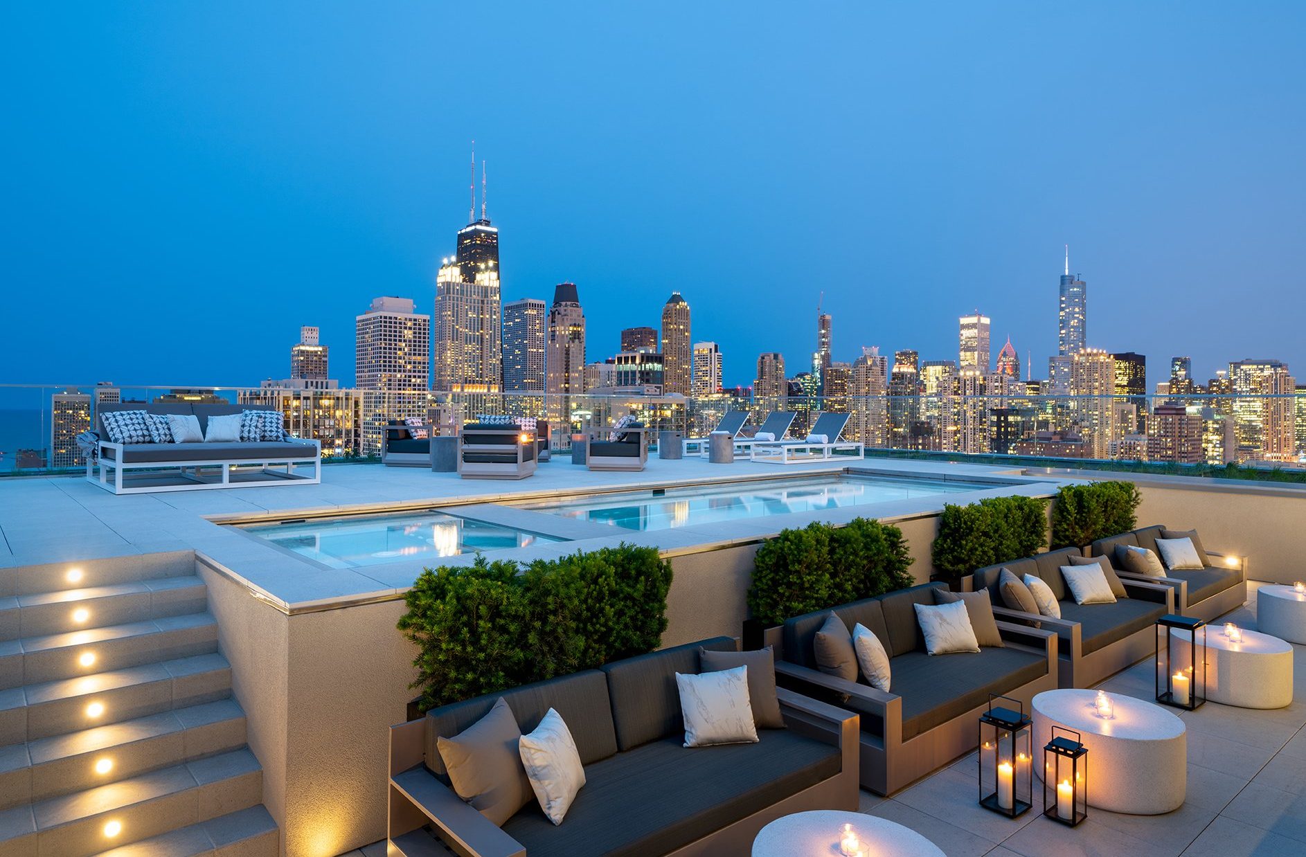 A modern rooftop lounge with stylish seating, glowing lanterns, and a pool overlooks a city skyline at dusk, with skyscrapers lit up against a blue evening sky.