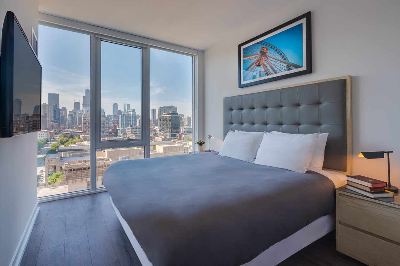 A modern bedroom with a large bed, gray headboard, bedside tables, and floor-to-ceiling windows offering a city skyline view. A framed Ferris wheel photo hangs on the wall above the bed.