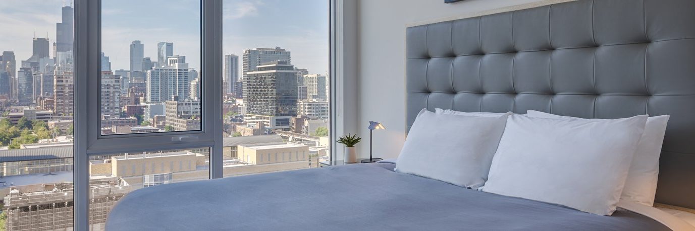A modern bedroom with a large gray upholstered bed, white pillows, and floor-to-ceiling windows offering a city skyline view during daytime. A small plant sits on a bedside table.