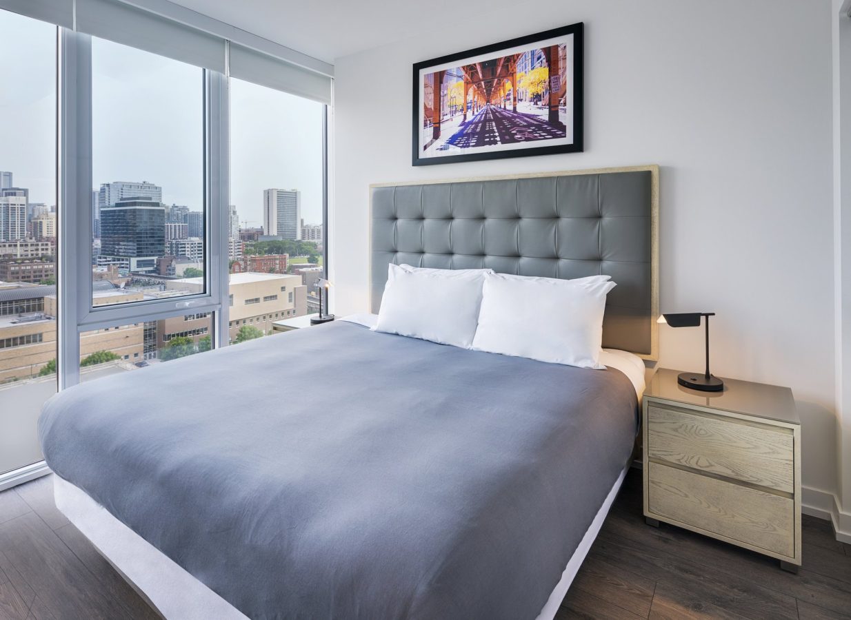 A modern bedroom with a large bed, gray tufted headboard, two white pillows, a wooden nightstand with a lamp, large windows overlooking a city skyline, and framed artwork on the wall above the bed.