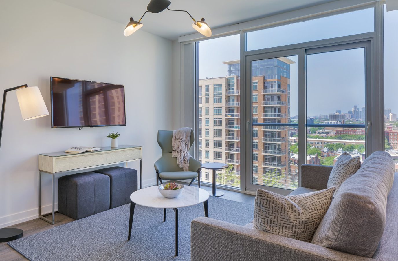 Modern living room with a gray sofa, patterned pillow, round coffee table, chair, TV on wall, desk, lamp, and large windows showing a cityscape with tall buildings and blue sky outside.