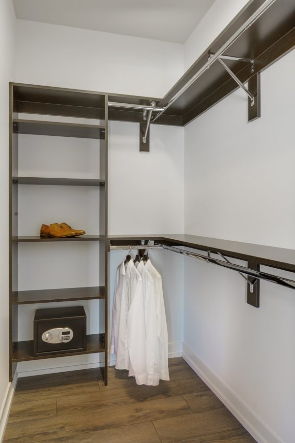 A modern, empty walk-in closet with dark shelves and hanging rods. Three white shirts hang neatly, a pair of brown shoes is on a shelf, and a safe is placed on the bottom shelf. The floor is dark wood.