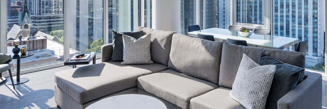 Modern high-rise living room with a gray sectional sofa and decorative pillows, a round white coffee table, and floor-to-ceiling windows offering city views. A dining table with chairs is visible in the background.