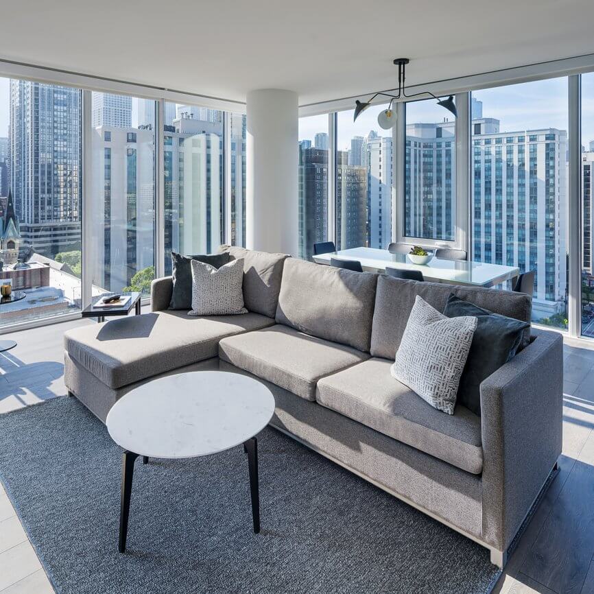 Modern living room with a gray sectional sofa, white marble coffee table, and large windows offering a city skyline view. Sunlight fills the space, which also includes a dining table in the background.