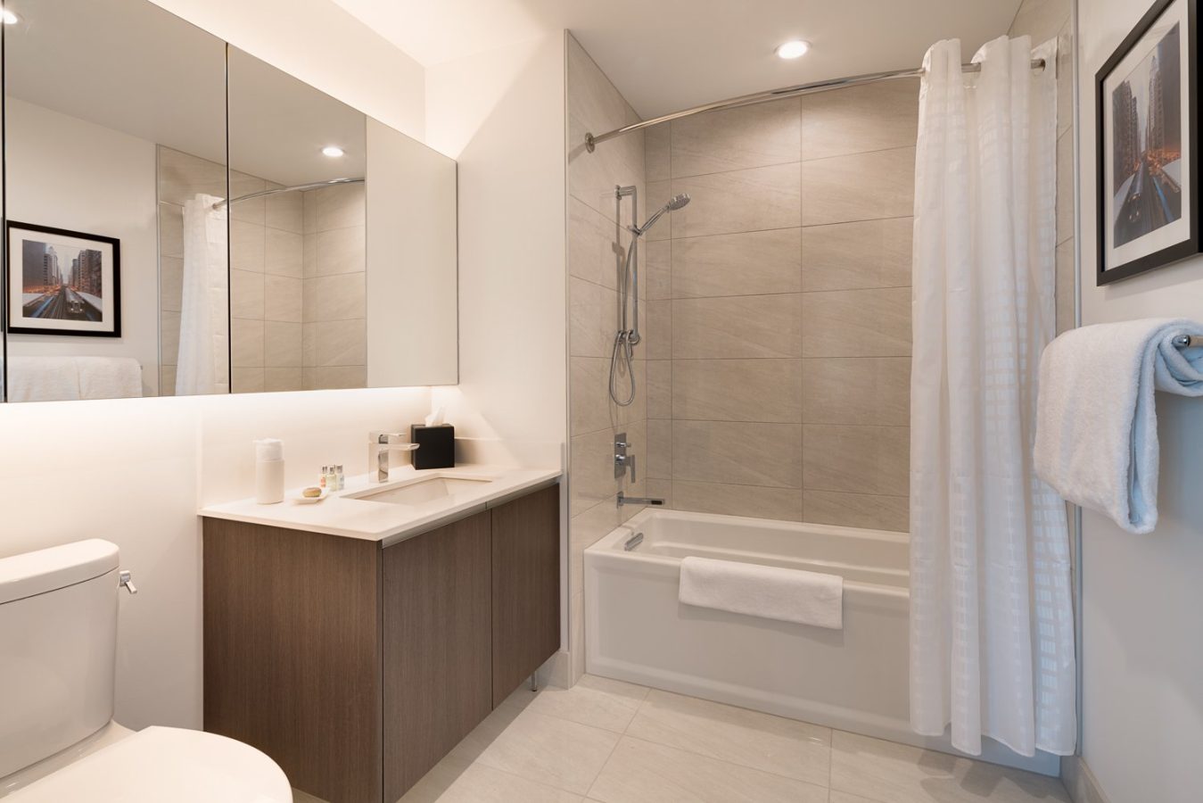 Modern bathroom with a bathtub and shower, white curtain, wall-mounted mirror cabinets, sink with toiletries, toilet, towel rack with a white towel, and framed photo on the wall. Neutral colors and bright lighting.