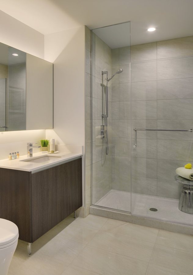 Modern bathroom with a large glass-enclosed shower, wall-mounted showerhead, gray tiled walls and floor, dark wood vanity with white countertop, mirror, and minimalistic decor.