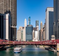 A white boat travels on the Chicago River beneath a red bridge, surrounded by tall skyscrapers under a clear blue sky in downtown Chicago.