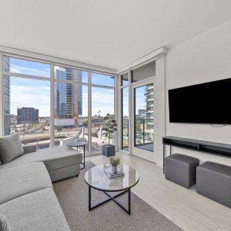 Modern living room with a gray sectional sofa, round coffee table, wall-mounted TV, and floor-to-ceiling windows offering a city view with tall buildings and blue sky.