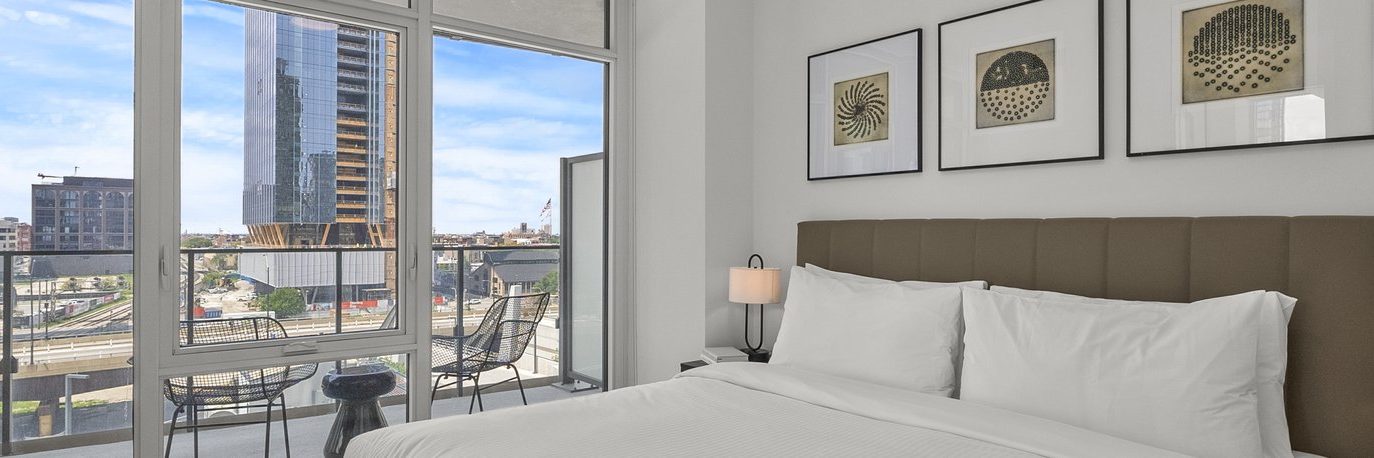 Modern bedroom with a large bed, three framed artworks on the wall, and a balcony with two chairs overlooking city buildings through floor-to-ceiling windows on a sunny day.