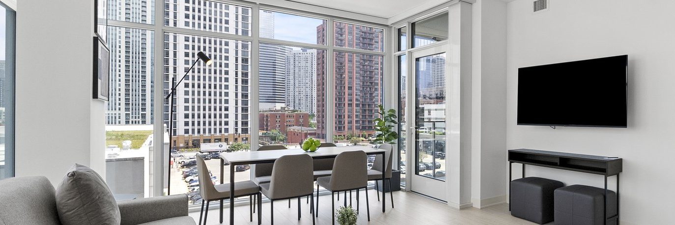 Modern apartment living room with large floor-to-ceiling windows, a dining table with four chairs, a wall-mounted TV, a gray sofa, and a city skyline view featuring tall buildings.