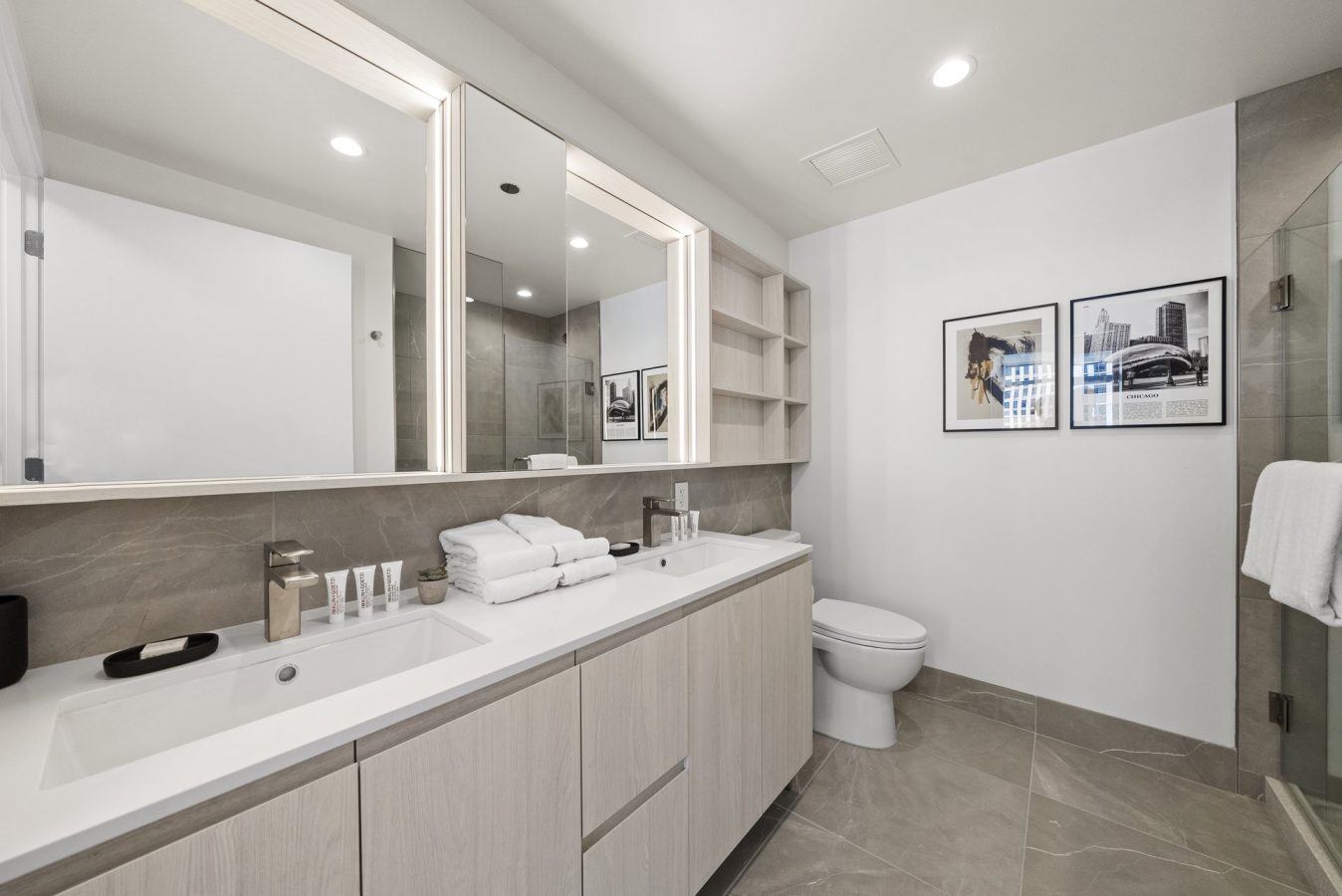 Modern bathroom with a double sink vanity, large mirrors, neatly folded towels, toiletries, wall art above the toilet, and a glass-enclosed shower; neutral tones and clean, contemporary design.