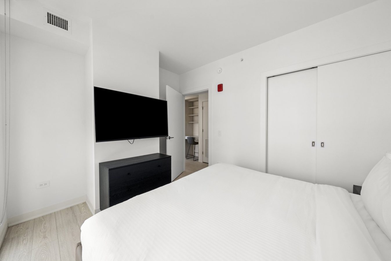 A minimal, modern bedroom with a white bed, a wall-mounted TV above a black dresser, sliding closet doors, and an open door revealing built-in shelves and a desk in a small adjacent room.