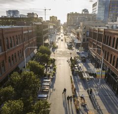 Aerial view of a city street lined with brick buildings, outdoor seating, and trees; a few people walk down the street, and sunlight casts long shadows across the scene.