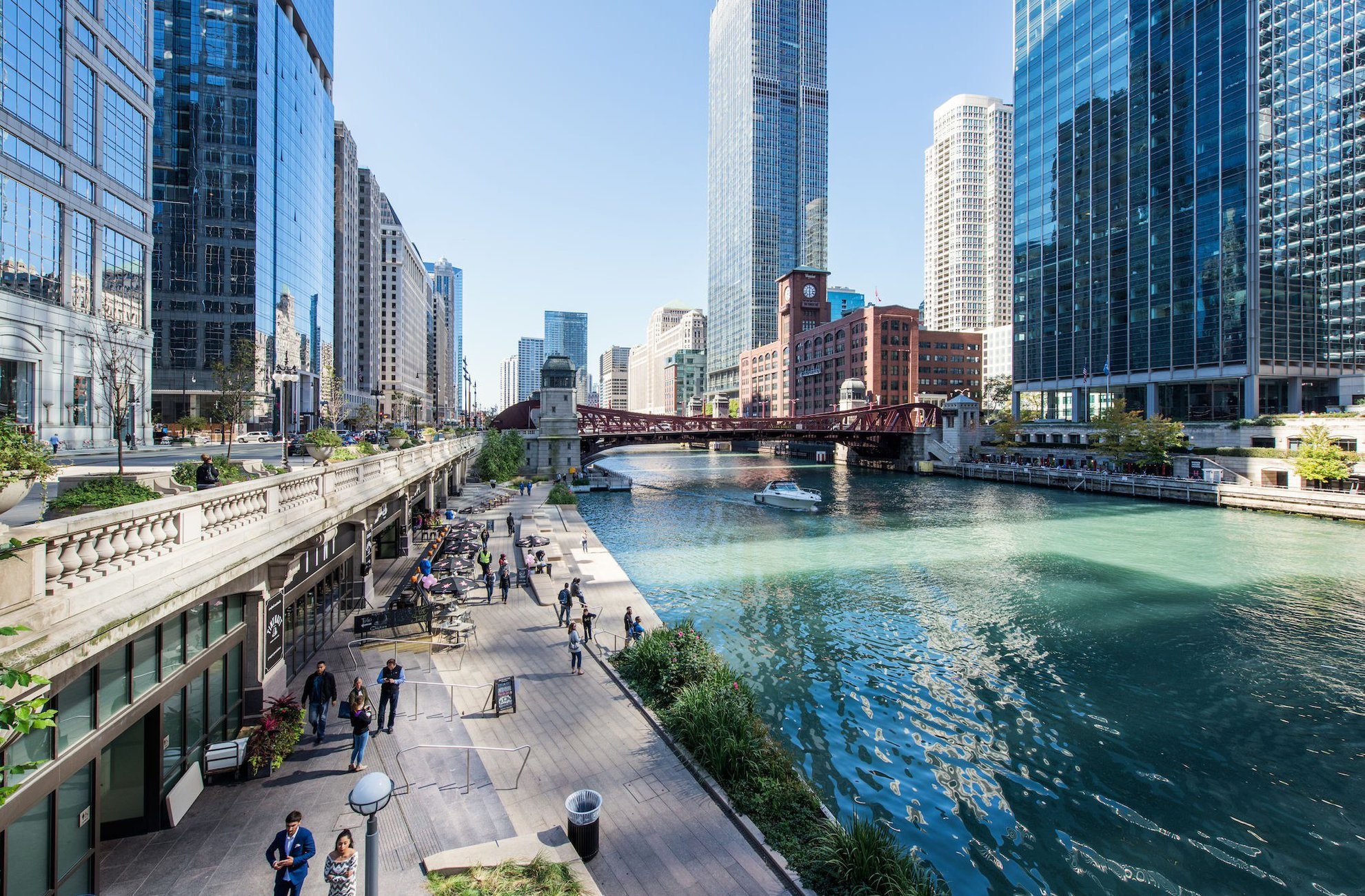 People walk and relax along a riverside promenade lined with plants and trees, with tall glass skyscrapers and a bridge over the river under a clear blue sky in a bustling city.