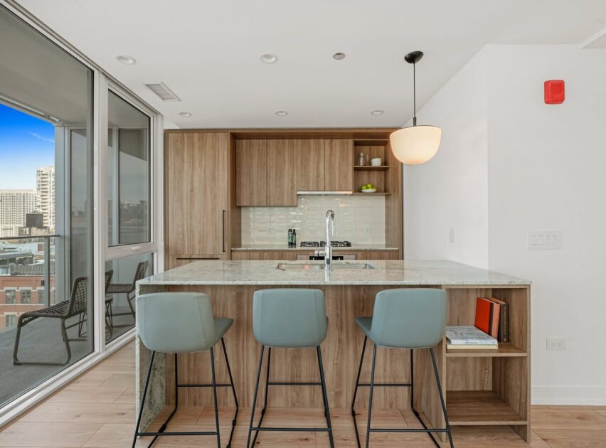 Modern kitchen with wooden cabinets, a marble island with three gray barstools, built-in shelves, and pendant lighting. There’s a balcony with city views visible through large glass doors on the left.
