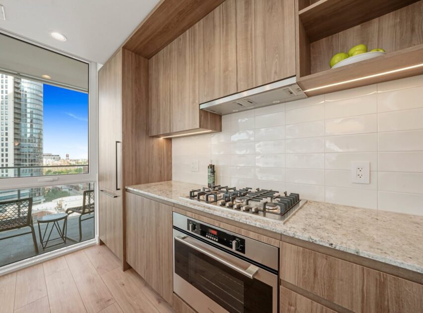 Modern kitchen with light wood cabinets, a built-in oven, and a gas stovetop. A large window opens to a balcony with outdoor seating and city views. White tile backsplash and marble countertop are visible.