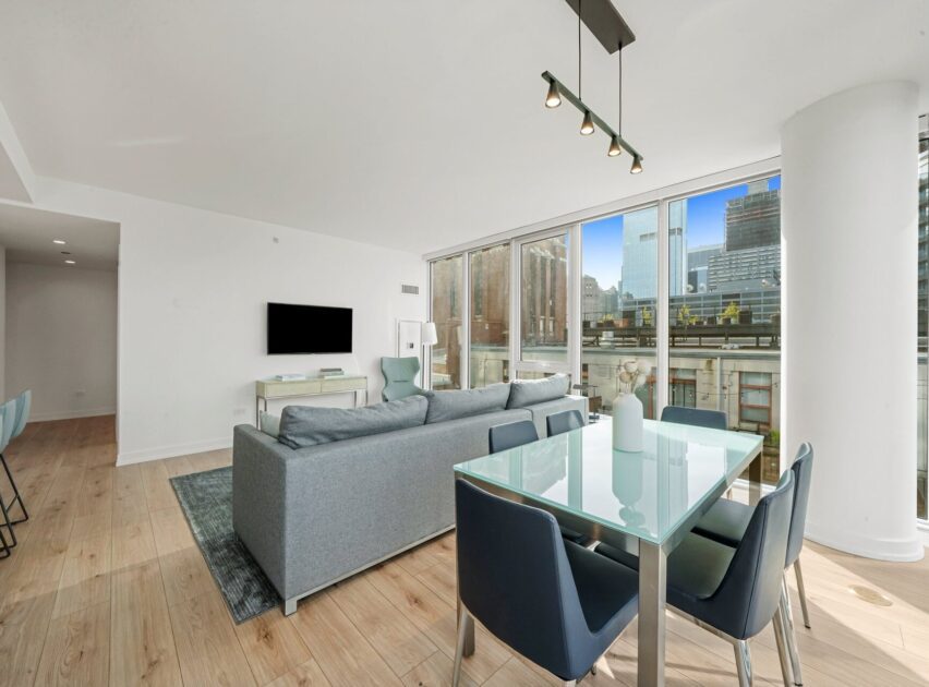 Modern living and dining area with light wood floors, a blue sofa, glass dining table with blue chairs, large windows offering a city view, and a mounted TV on a white wall.