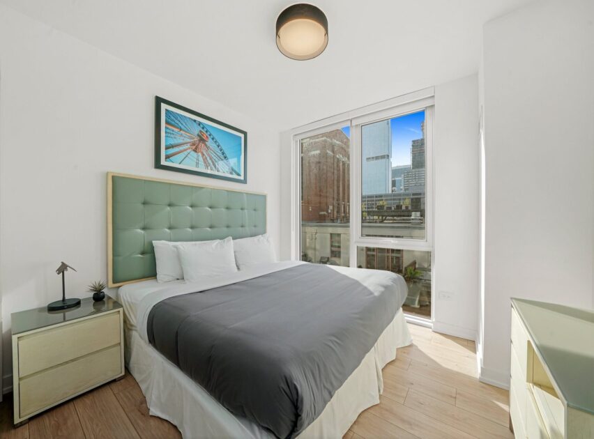 Bright, modern bedroom with a large bed, padded headboard, two nightstands, and a cityscape view through floor-to-ceiling windows. A framed artwork hangs above the bed.