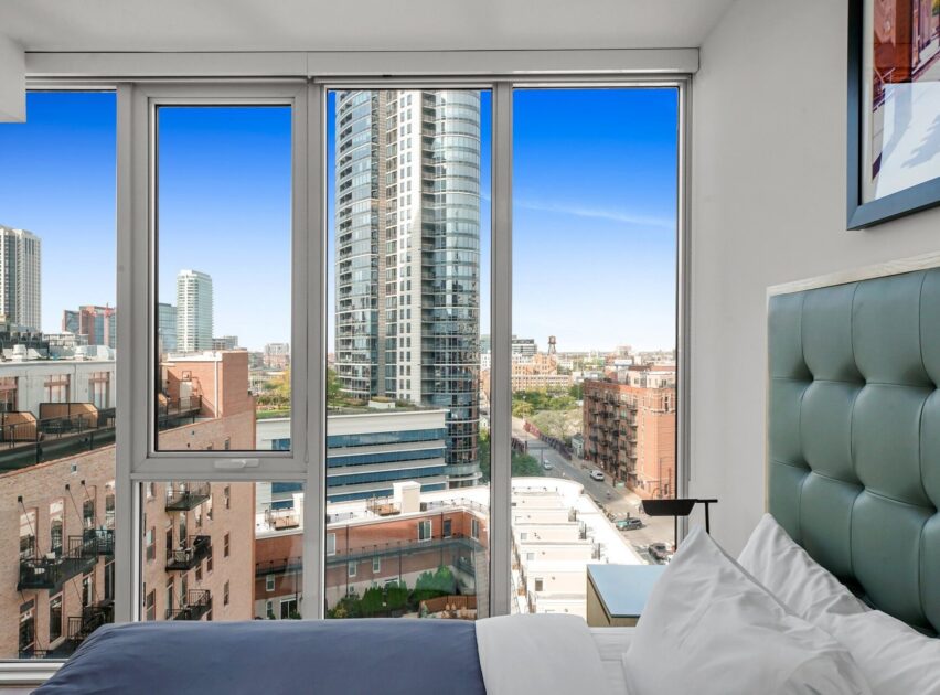 Modern bedroom with floor-to-ceiling windows offering a city view of tall buildings, a clear blue sky, and urban streets below. Part of a bed with white pillows is visible in the foreground.