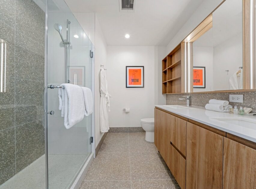 Modern bathroom with a glass shower, wood cabinets, double sinks, wall-mounted mirror, two framed orange prints, and a robe hanging by the toilet. White towels are neatly arranged throughout the space.