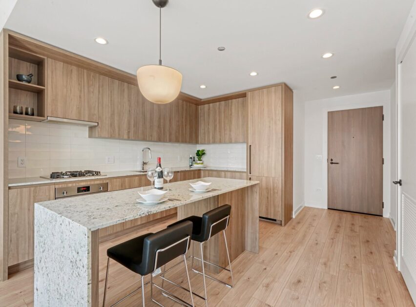 Modern kitchen with light wood cabinets, white tile backsplash, marble island with two place settings, black bar stools, and stainless steel appliances. Pendant light hangs above island on natural wood floor. Door visible in background.