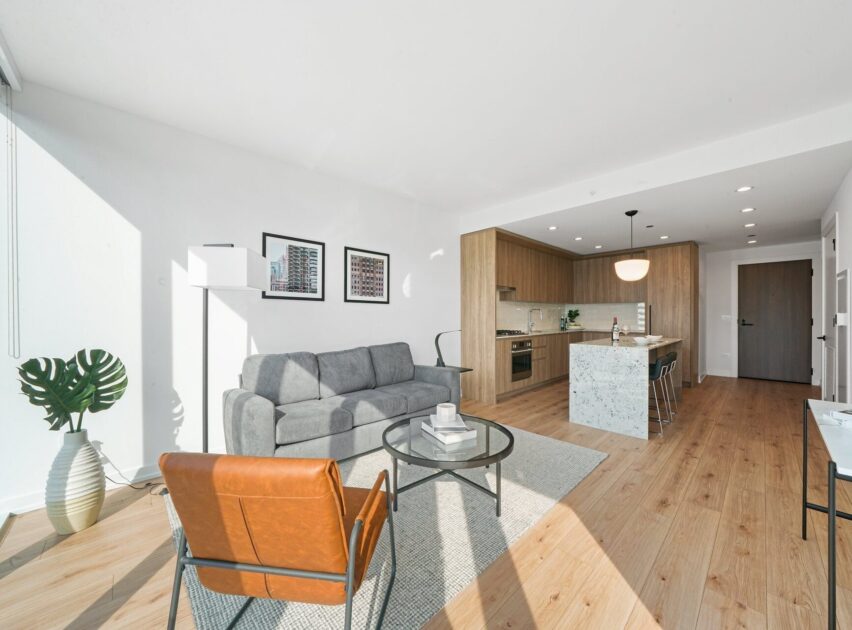 Bright, modern apartment interior with a gray sofa, brown leather chair, glass coffee table, large plant, and open kitchen featuring wooden cabinets and a marble island. Natural light fills the space through large windows.