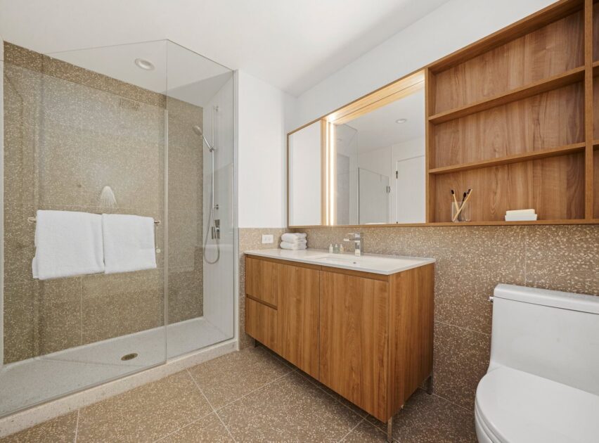 Modern bathroom with a glass-enclosed shower, wooden vanity with sink, large mirror, open wooden shelves, and a white toilet. Towels are hanging inside the shower. The decor is minimalist and neutral-toned.