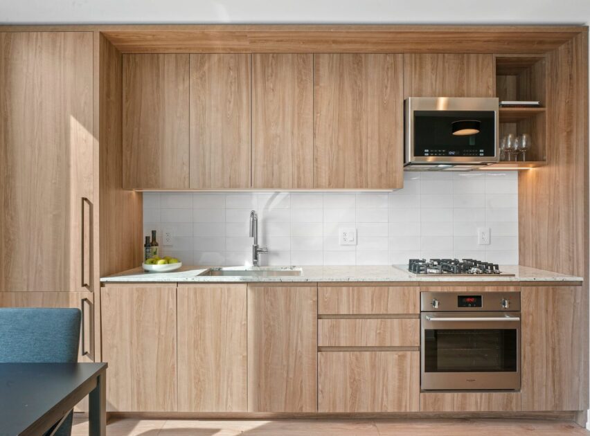 Modern kitchen with light wood cabinets, white tile backsplash, built-in oven, gas stovetop, stainless steel microwave, sink, and countertop with fruit bowl and utensils. A dark dining table and blue chair are partially visible.