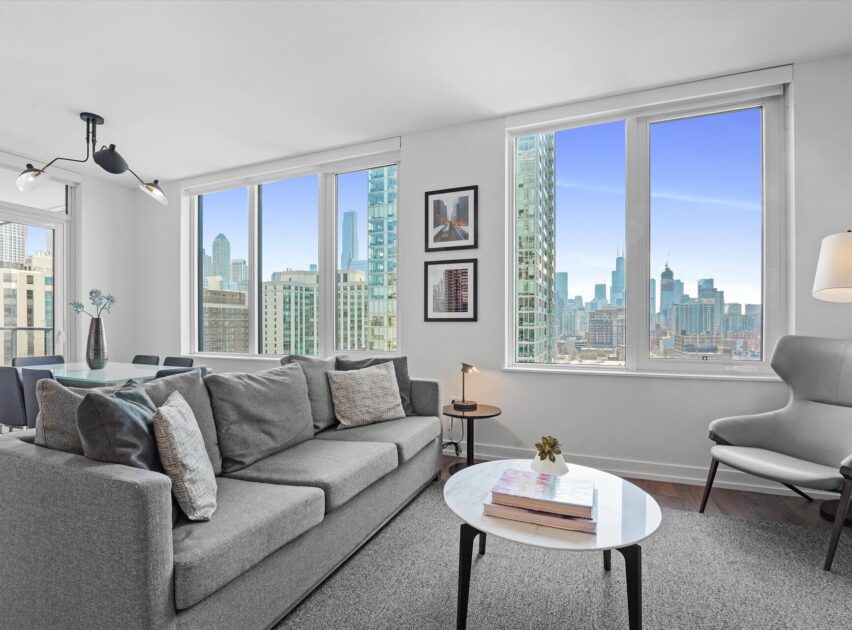 A modern living room with a gray sofa, coffee table, and armchair, large windows offering a city skyline view, and contemporary decor with framed art on white walls.