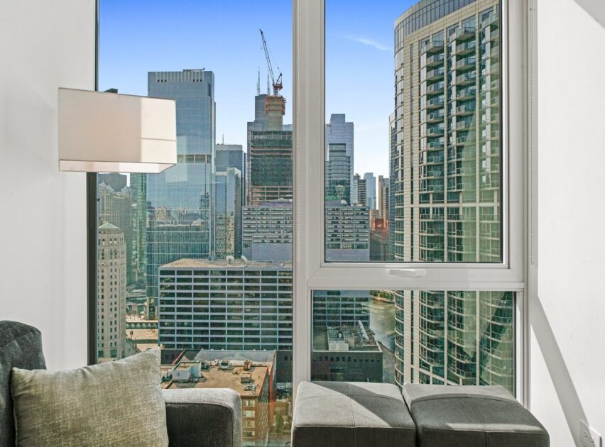 A modern high-rise apartment with floor-to-ceiling windows overlooking a cityscape of tall buildings, construction cranes, and a clear blue sky; a gray sofa and ottoman are visible in the foreground.