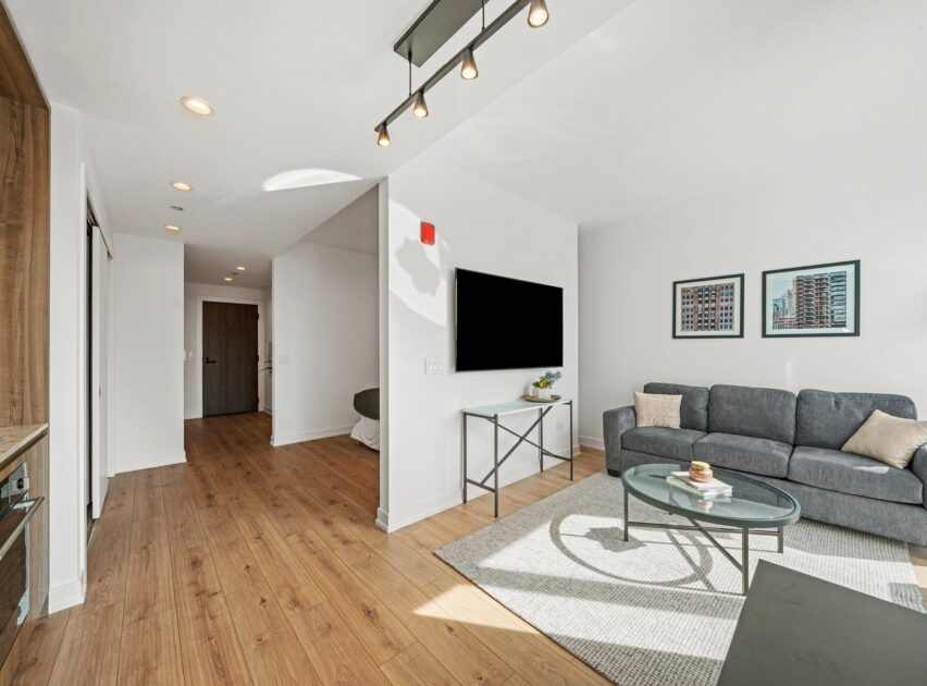 Modern living room with light wood floors, gray sofa, glass coffee table, wall-mounted TV, and framed cityscape art. Bright space with natural light and recessed ceiling lights. Hallway leads to a wooden door.