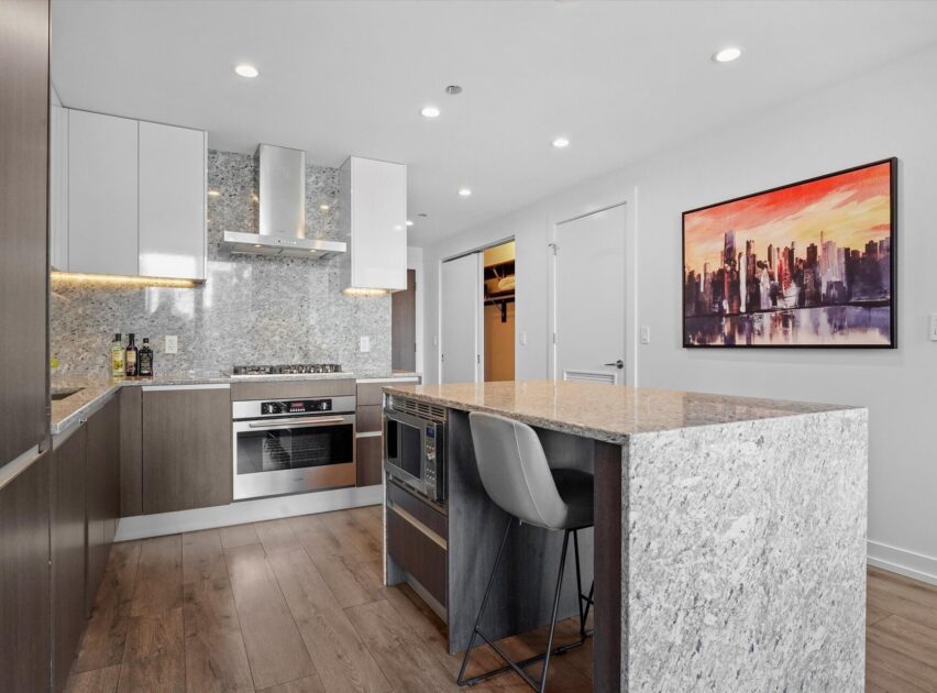 Modern kitchen with granite countertops, stainless steel appliances, wood flooring, and a gray island with a built-in oven and a barstool. A colorful cityscape painting hangs on the white wall.