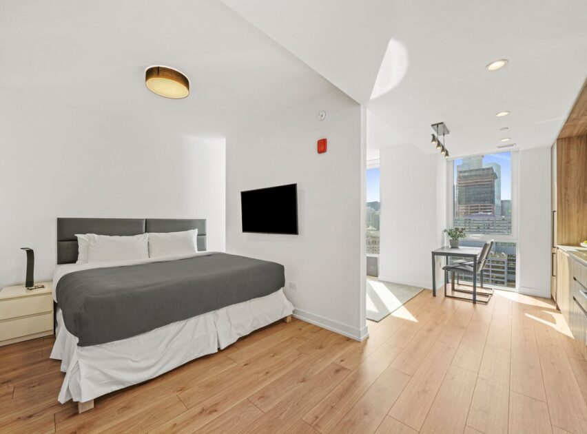 A modern, bright studio apartment features a neatly made bed with gray bedding, wood flooring, a wall-mounted TV, and large windows offering a city view near a small dining area.
