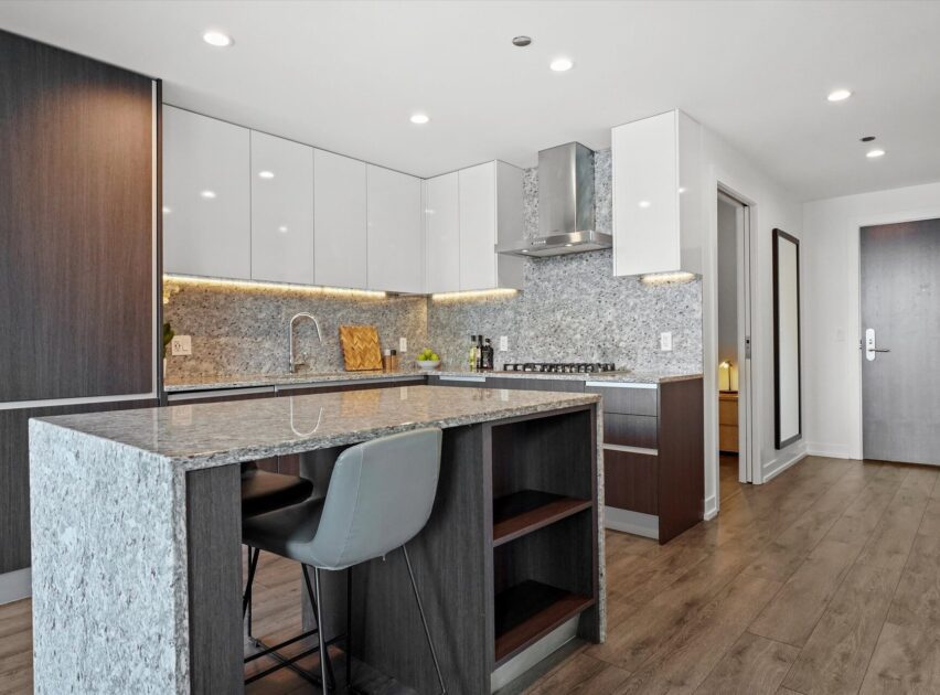 Modern kitchen with gray granite countertops, wood and white cabinets, built-in stove, and integrated oven. An island with a cushioned gray chair stands on a wood floor, with recessed lighting and contemporary decor.