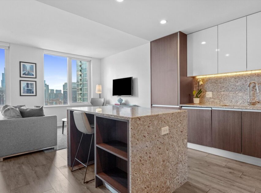 Modern apartment with an open kitchen featuring granite countertops, a built-in sink, and wood cabinets. A living area with a sofa and TV sits by large windows overlooking a cityscape with tall buildings.