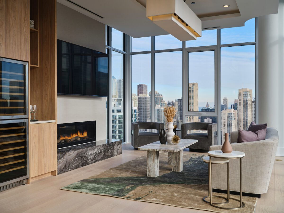 A modern living room with floor-to-ceiling windows showing a city skyline, a marble coffee table, neutral-toned furniture, a built-in fireplace, a wine fridge, and decorative vases on the table and side table.