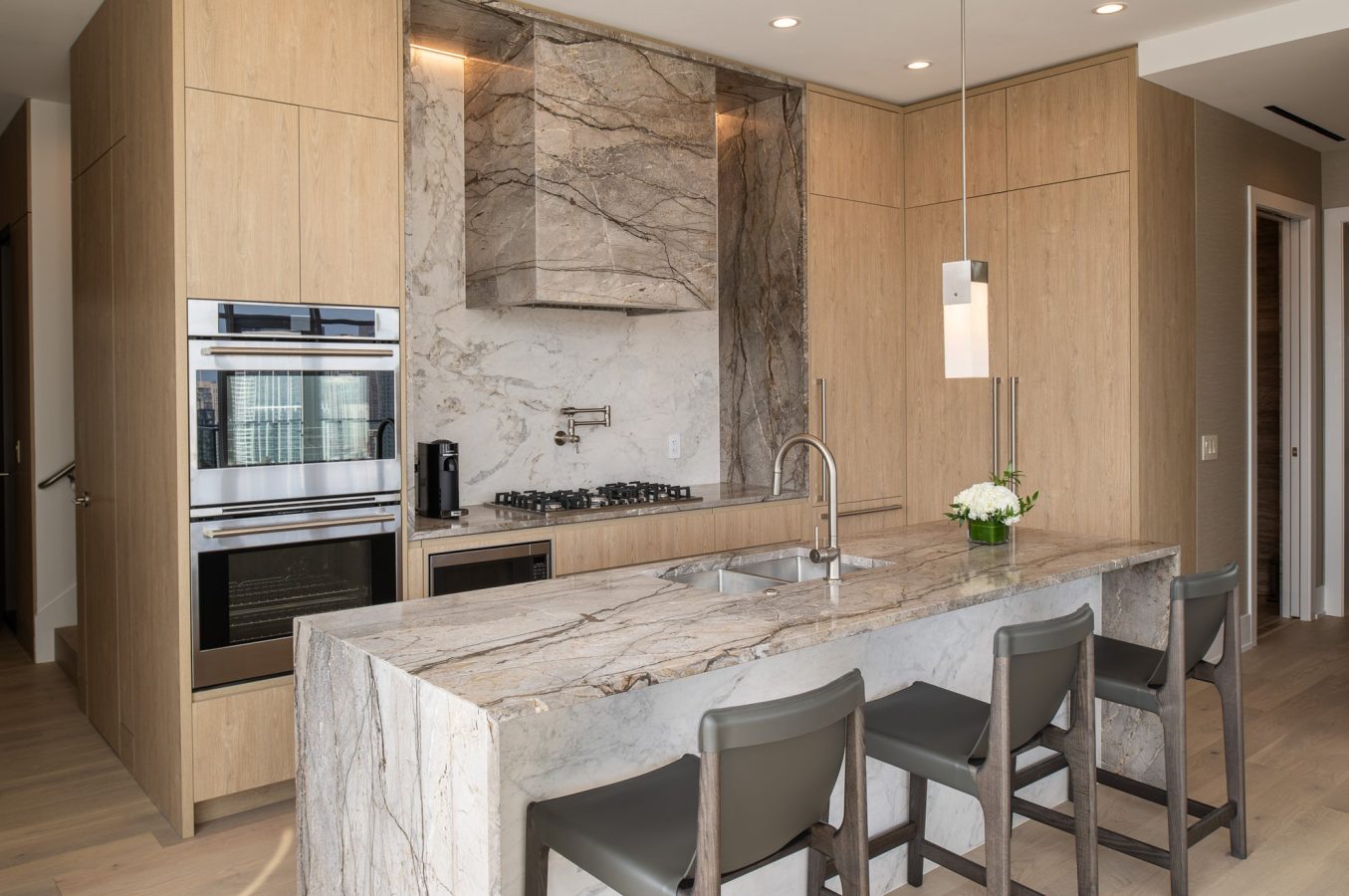 Modern kitchen with light wood cabinets, marble backsplash and countertops, built-in stainless steel oven, stovetop, sink on island, three gray bar stools, and a pendant light above a vase of white flowers.