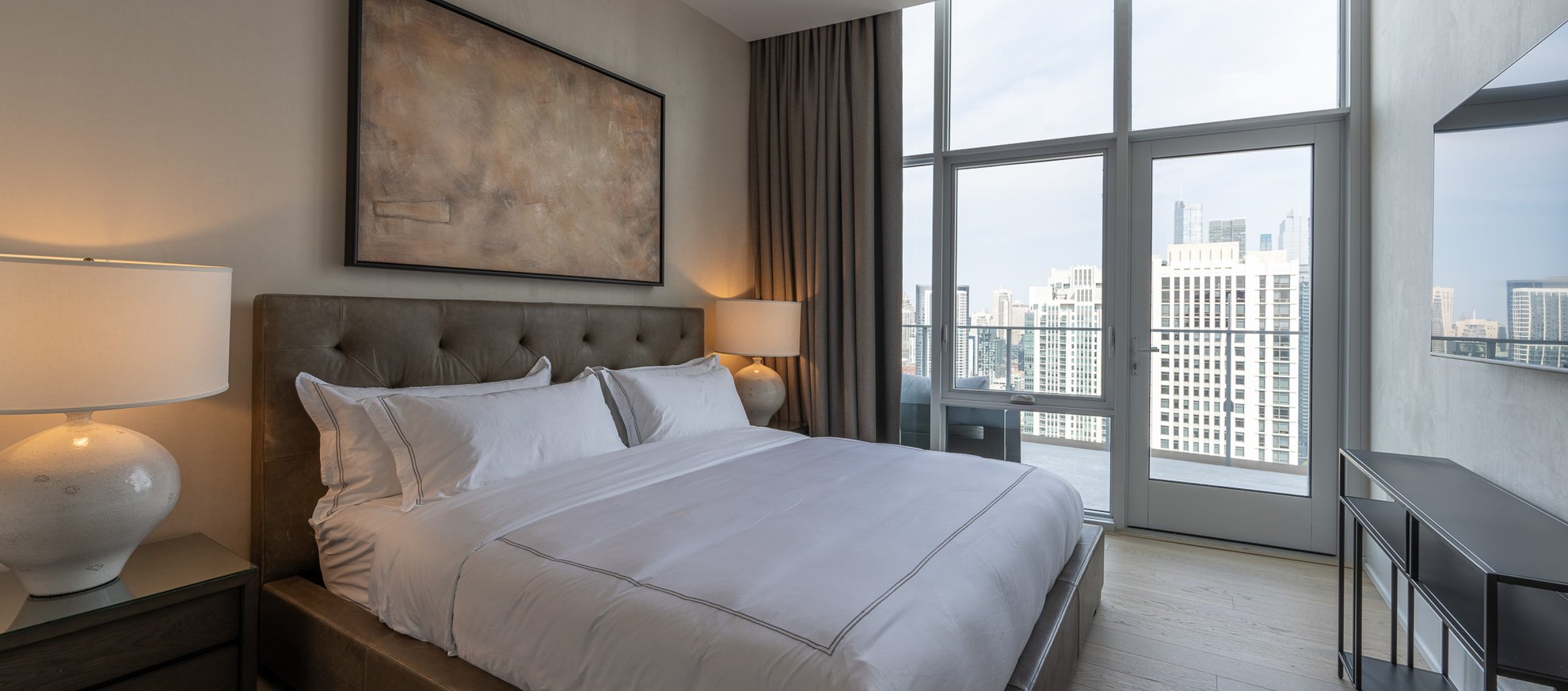 A modern bedroom with a large bed, white bedding, two bedside lamps, a wall-mounted TV, and floor-to-ceiling windows offering a city view with tall buildings.