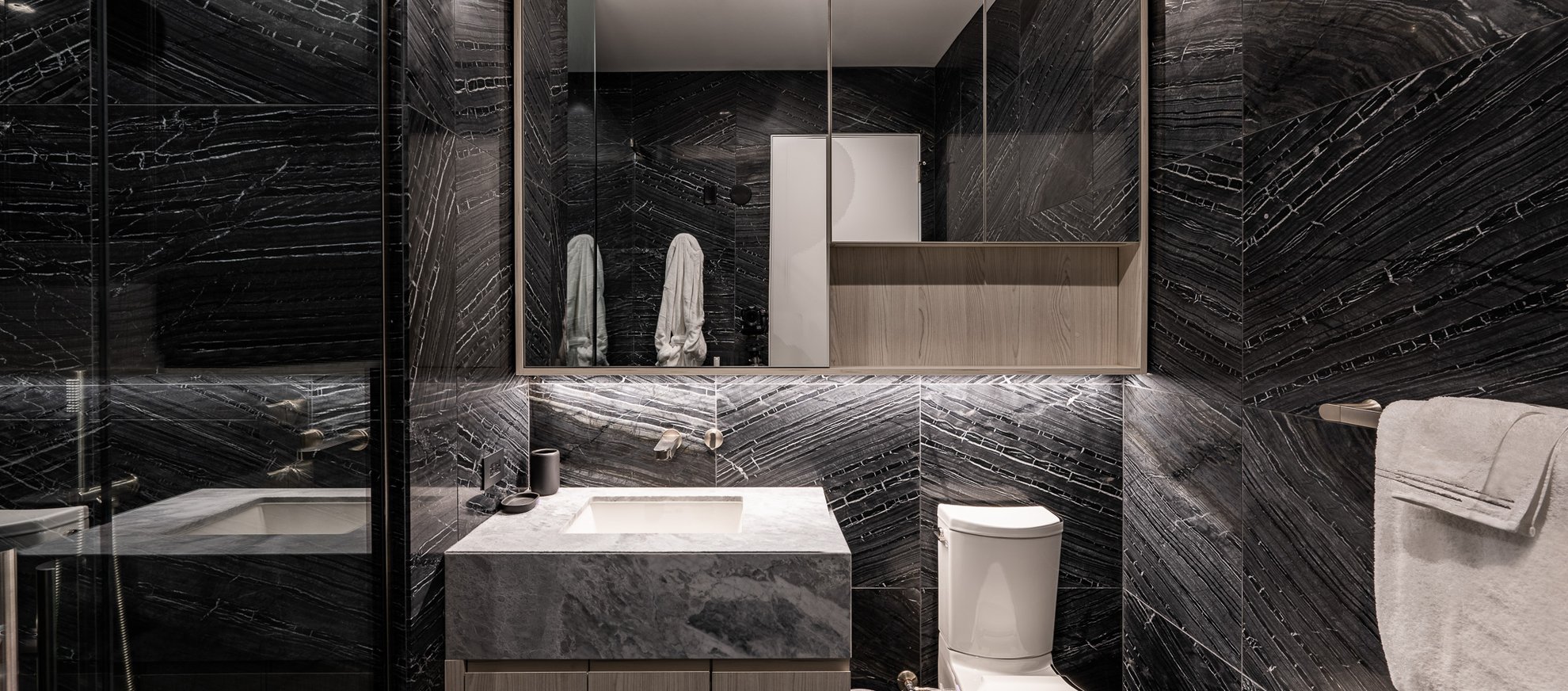 Modern bathroom with dark, marble-patterned walls, a large mirror, a white toilet, a sink with a marble countertop, towels hanging on racks, and built-in shelves above the sink.