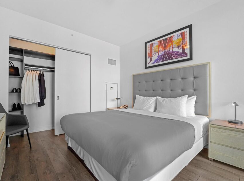 Modern hotel room with a large bed, gray headboard, two nightstands, table lamps, open closet with clothes, wood flooring, and a colorful city artwork above the bed. The room is bright and neatly organized.