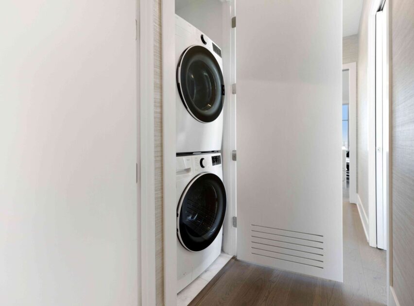 A modern laundry closet with a stacked white washer and dryer, partially concealed behind a white door, is shown in a bright, minimalist hallway with wood flooring.